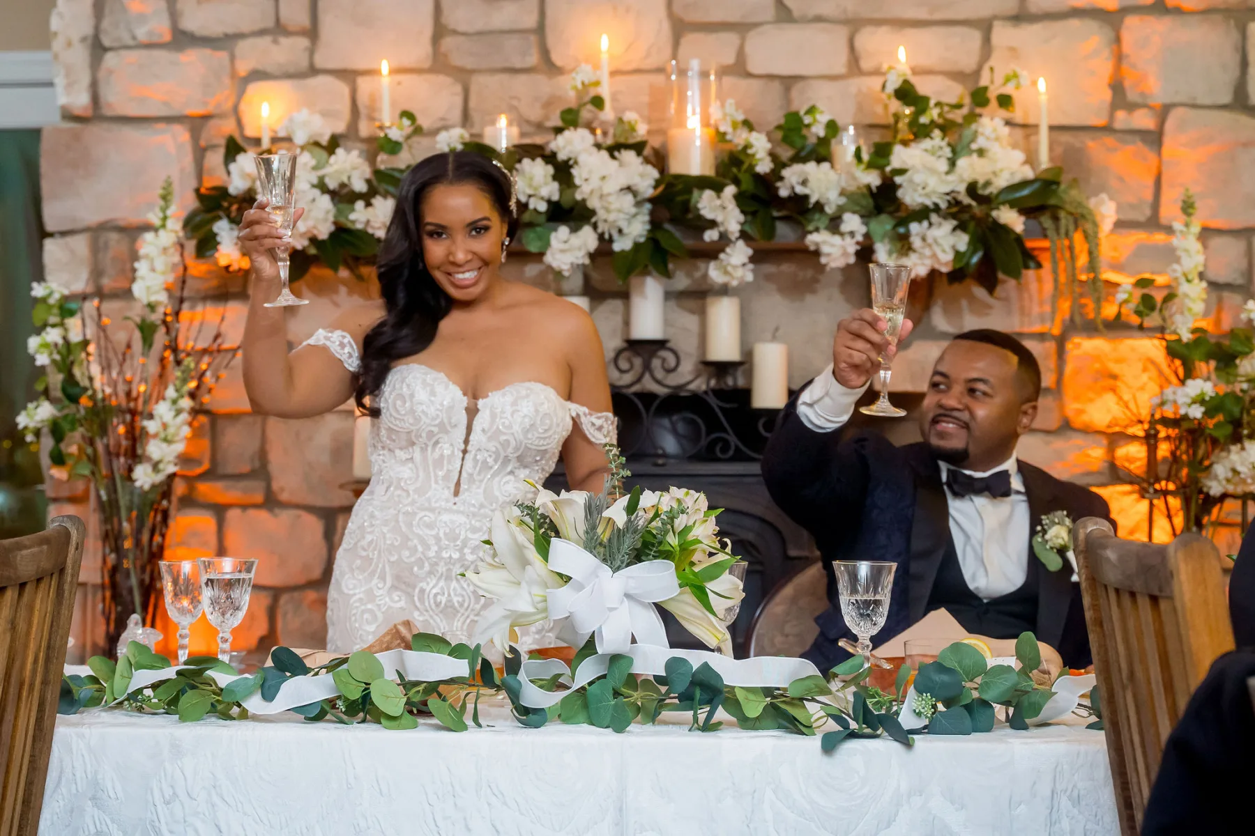 A bride and groom raising champagne flutes in a toast at a candlelit reception, eucalyptus garland and ivory florals on the sweetheart table.